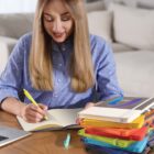 A woman with long blonde hair is sitting at a table, smiling and writing in a notebook. She is wearing a blue polka dot shirt. A stack of colorful notebooks and a highlighter are on the table beside her. A sofa is in the background.