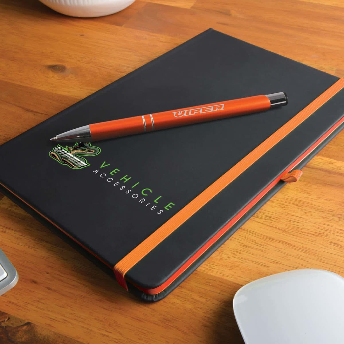 A black notebook with Vehicle Accessories written in green lies on a wooden table. An orange elastic band secures it, and a matching orange pen labeled Viper rests on top. A portion of a white object is visible at the bottom right.