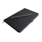A closed black notebook with an elastic band lays on a surface with a black pen resting on top. The notebook has a ribbon bookmark peeking out from the right side.