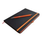 A closed black notebook with an orange elastic band lies on a flat surface. An orange and silver pen rests diagonally on top of the notebook. An orange ribbon bookmark is visible.