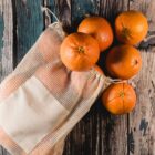 A netted bag filled with six oranges spills onto a rustic wooden surface. The oranges are bright and fresh, and the wood features a weathered texture with shades of brown and blue.