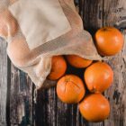 A mesh bag, partially open, displays several ripe oranges spilling out onto a rustic wooden surface. The bag has a plain fabric label. The oranges are bright and glossy, contrasting with the dark wood background.