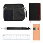 A flat lay of office supplies includes a black zippered pouch, a black notebook with a red stripe, a black pen, a set of colored pencils in a brown box, and a white 15 cm ruler.