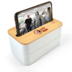 A smartphone is placed on a light wooden box with a video playing, showing three people. The box has the logo MR gadget on it. The setup suggests an amplified sound system for the phone.