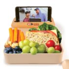 A bento box with a sandwich, green grapes, cherry tomatoes, blueberries, carrots, almonds, and walnuts. A phone playing a video is propped up in the lid. The lid has MR SUSHI and a sushi logo on it.