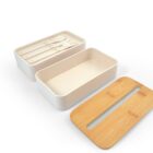 A three-piece lunch box set with a bamboo lid, a beige container, and a top tray holding a fork, spoon, and knife. The tray fits inside the container, and the lid has two long slits. The design is minimalist.
