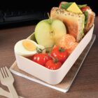 A beige lunchbox containing half a green apple, a boiled egg, cherry tomatoes, a peeled mandarin, and a sandwich with lettuce and cheese. It is placed on a wooden surface with a fork and knife beside it.