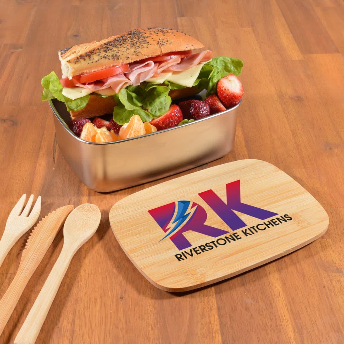 A sandwich with lettuce, ham, cheese, and tomato sits in a metal lunchbox, accompanied by strawberries and orange slices. The wooden lid, placed nearby, is branded with RK Riverstone Kitchens. Bamboo cutlery is arranged beside it on a wooden table.