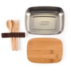 A stainless steel lunchbox with a wooden lid sits next to a set of wooden utensils, including a spoon, fork, and knife, held together by a brown strap.