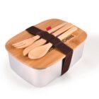A rectangular lunchbox with a wooden lid secured by a dark strap. Wooden utensils, a fork, spoon, and knife, are attached on top of the lid. The lid has a logo and text partially visible. The box itself is metallic.