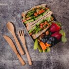 Lunchbox containing vegetable sandwiches, strawberries, blueberries, almonds, carrot, and celery sticks, with wooden cutlery set on a textured gray surface.