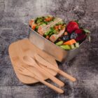 A stainless steel lunchbox containing a sandwich with lettuce, tomato, and possibly turkey, alongside strawberries, blueberries, and sliced carrots and cucumbers. Wooden cutlery and a small board are placed beside it on a textured surface.