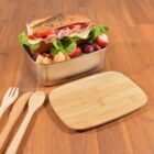 Lunchbox with a sandwich, lettuce, ham, cheese, strawberries, and orange slices on a wooden table. The lid is propped up beside the box. Wooden utensils are placed to the left of the box.