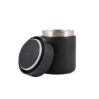 A black insulated container with a stainless steel interior and a detachable lid sits open. The lid rests in front, showing a sealing ring designed to keep contents secure and temperature stable.