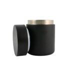 A black cylindrical jar with a detached lid. The jar has a metallic top rim and a matte finish. The lid is leaning against the jar, revealing the open top.