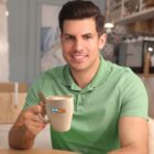 Man in a light green polo shirt holds a mug with the One Tech logo, smiling, in a cozy room with blurred background elements including a painting and hanging light fixture.