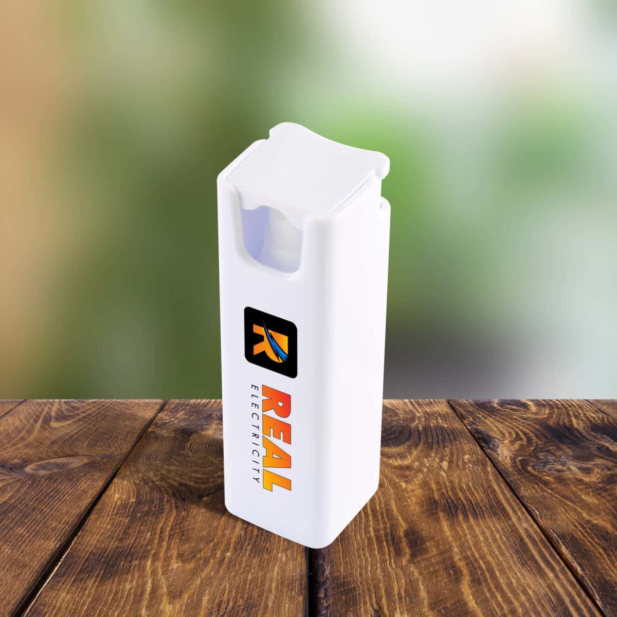 A white, rectangular, reusable lighter with the Real Electricity logo on the side, featuring a large R and the company name below it. It rests on a wooden surface with a blurred green background.