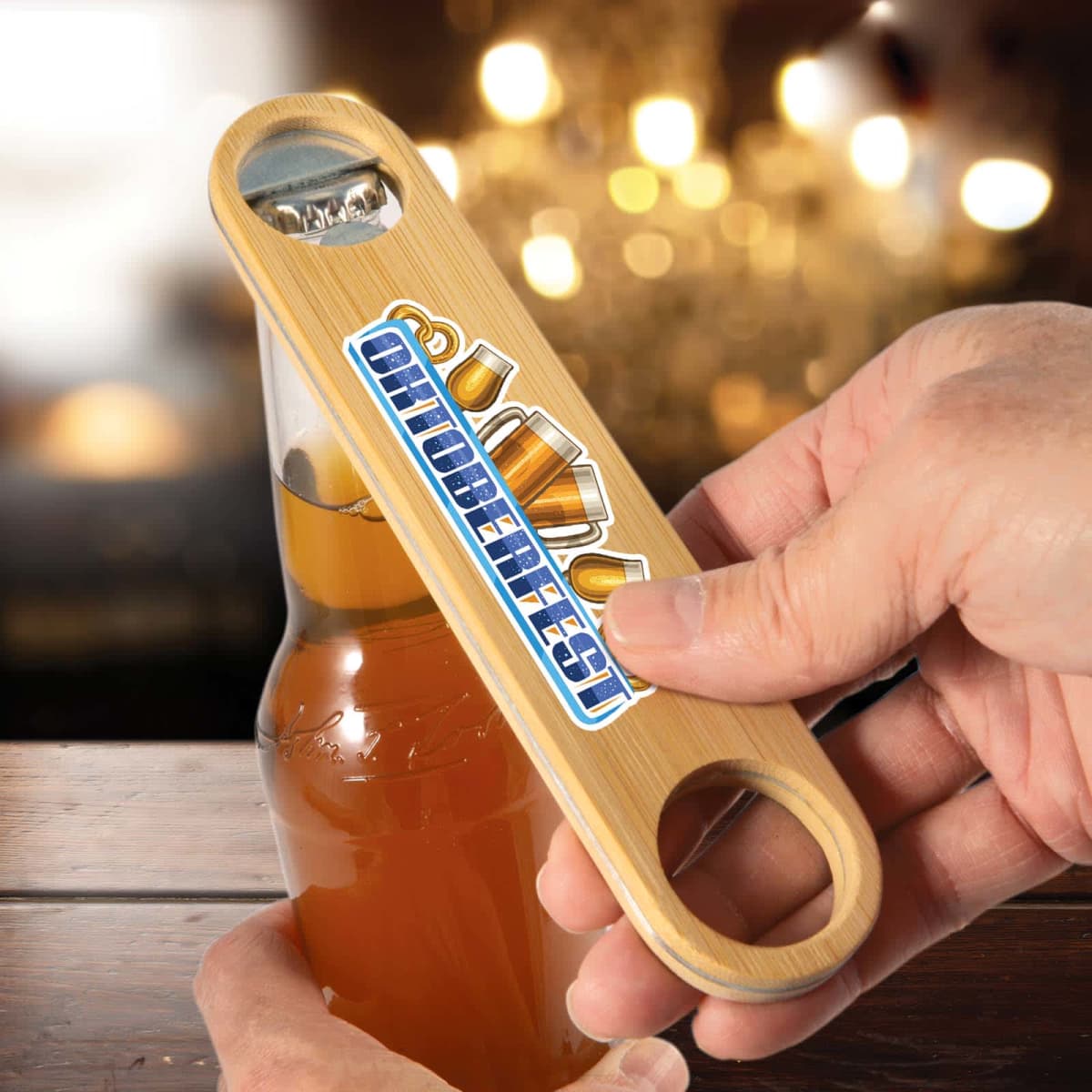 Close-up of a person using a wooden bottle opener with Oktoberfest and beer mug graphics to open a beer bottle. The setting is dimly lit, with a blurred background hinting at a festive atmosphere.
