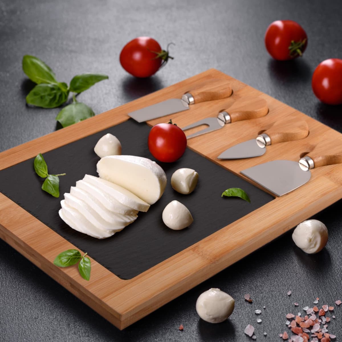 A wooden cheese board with black slate displays sliced mozzarella, whole mozzarella balls, and a cherry tomato. Cheese knives are arranged on the board. Basil leaves and cherry tomatoes are scattered around. Pink salt crystals are on the black surface.