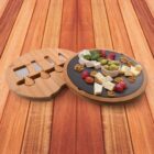 A round bamboo cheese board with a slate top holds assorted cheeses, grapes, olives, walnuts, and herbs. Next to it is a separate bamboo tray with cheese knives. The board is placed on a wooden surface.