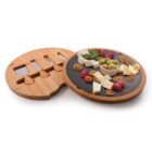 A round bamboo cheese board with a pull-out drawer displaying cheese knives. The top surface holds assorted cheeses, grapes, walnuts, and olives arranged around a brie wheel, on a black slate center.
