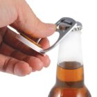 A hand using a bottle opener to open a brown glass bottle with a metal cap. The liquid inside is amber-colored.