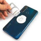 A hand is attaching a magnetic car mount holder to the back of a teal smartphone. The phone has a dual camera and a logo on the bottom left corner. The holder is silver and white, with a circular shape.