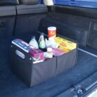 A black car trunk organizer containing various grocery items, including beverage bottles, a cereal box, a pack of mixed nuts, cleaning wipes, and other packaged foods. The organizer is placed on the carpeted floor of a vehicles trunk.