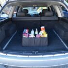 Open trunk of a car with a black organizer containing various grocery items, such as bottled drinks, cereal, canned goods, and snacks. The car is parked outdoors and sunlight is casting shadows inside the trunk.