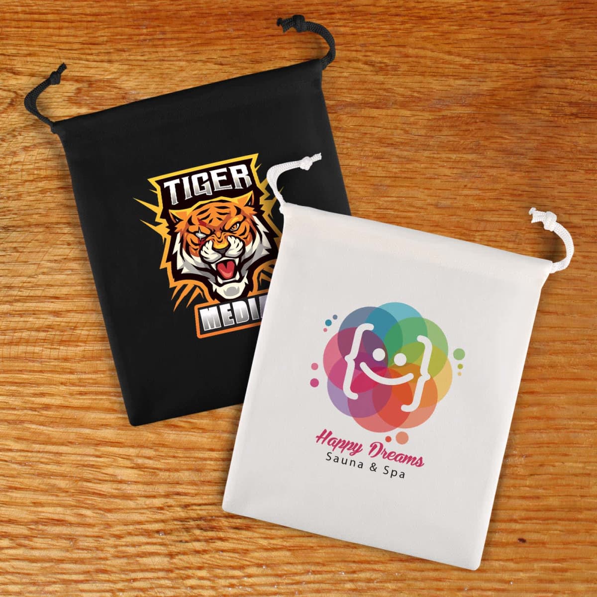 Two drawstring bags on a wooden surface: the black bag features a fierce tiger design with the words Tiger Media, and the white bag displays a colorful logo with the text Happy Dreams Sauna & Spa.
