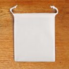 A small white cloth drawstring bag lies flat on a wooden surface. The bag has two white strings at the top for closing and opening.