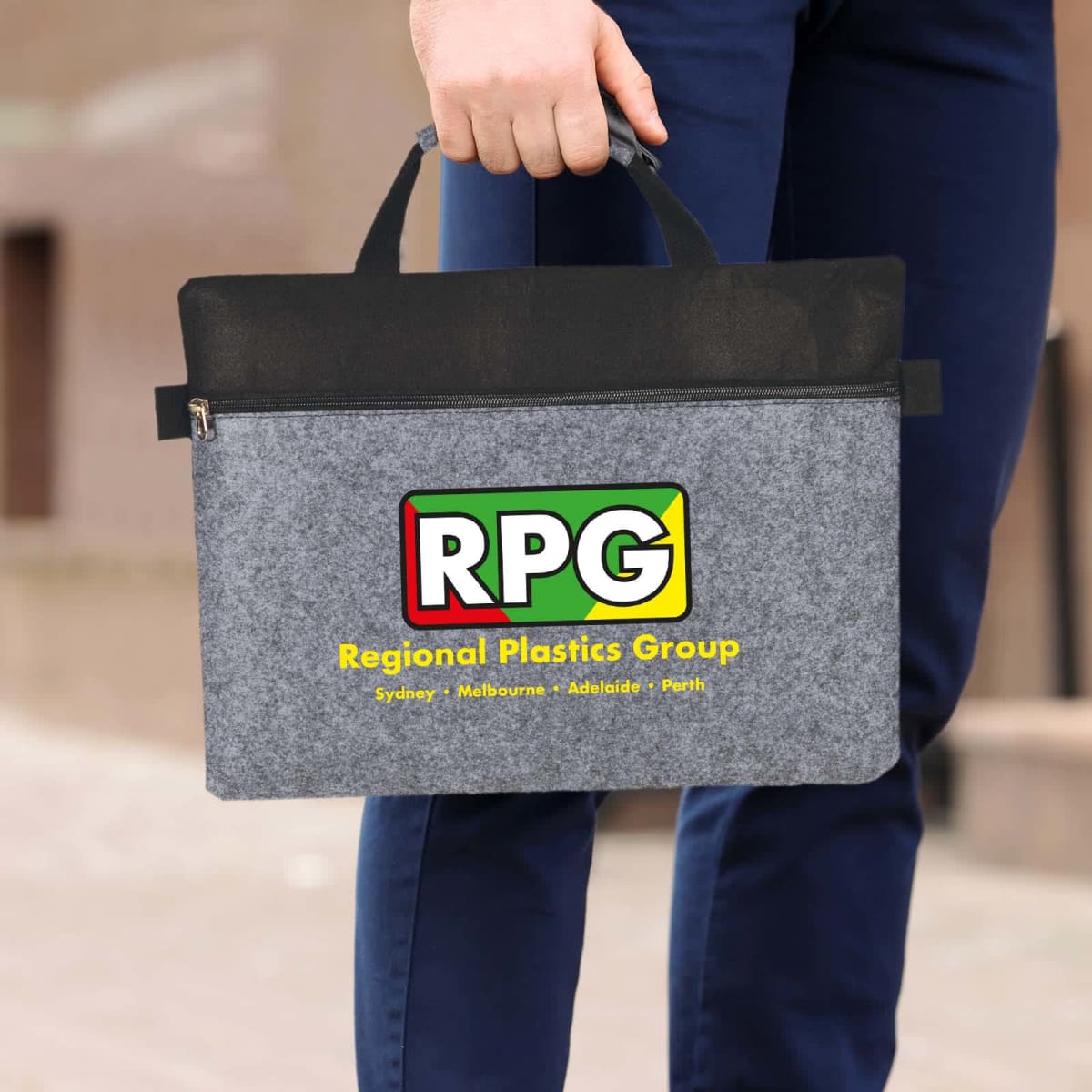 Person holding a gray and black tote bag with RPG and Regional Plastics Group printed on it, alongside the cities Sydney, Melbourne, Adelaide, and Perth. The person is wearing blue pants and a brown background is visible.