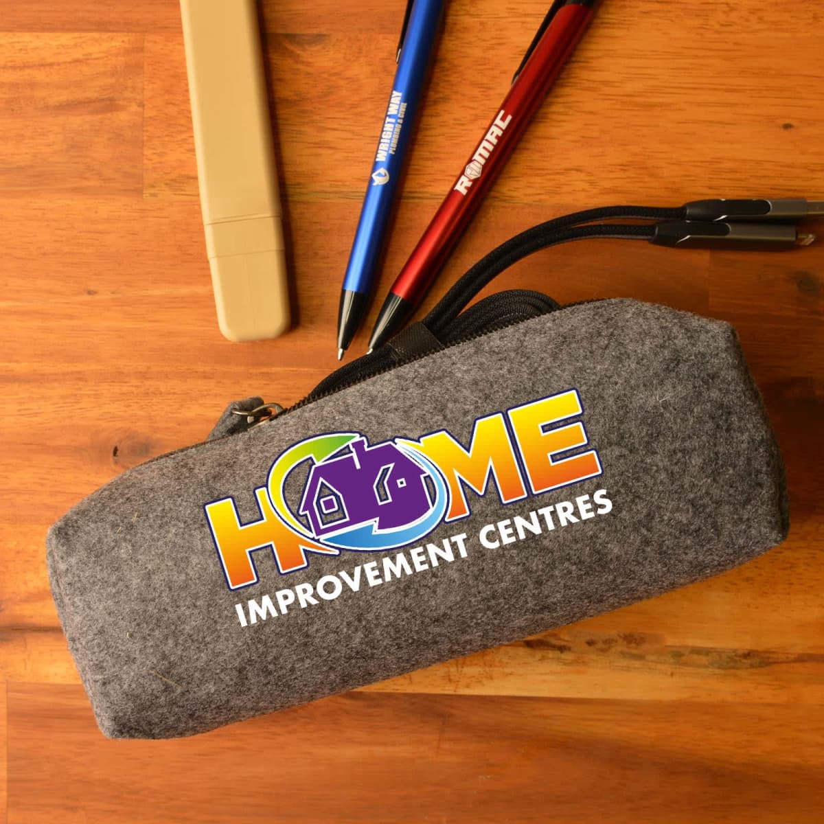 A felt pencil case with the Home Improvement Centres logo is on a wooden table. Nearby are beige, blue, and red pens, each with FINE TEC written on them. The case zipper is partially open, revealing pen clips inside.