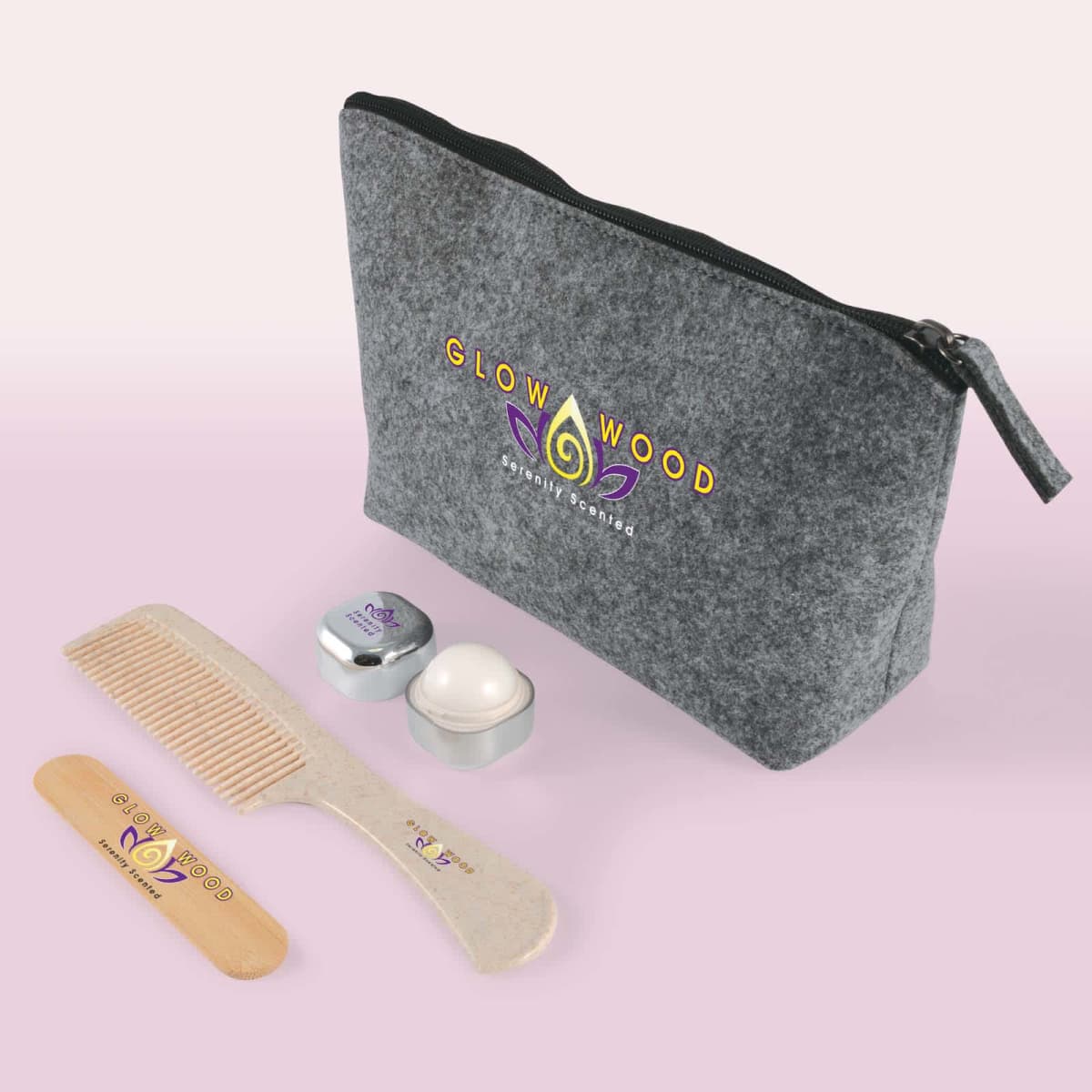 A travel set with a gray zippered pouch labeled Glow Wood, a wooden comb, a small metallic jar, and a round container. The items are arranged on a light pink surface.