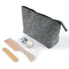 A gray zippered pouch is placed next to a small round metal container, a bamboo comb, and a wooden stick. The items are arranged neatly against a white background.