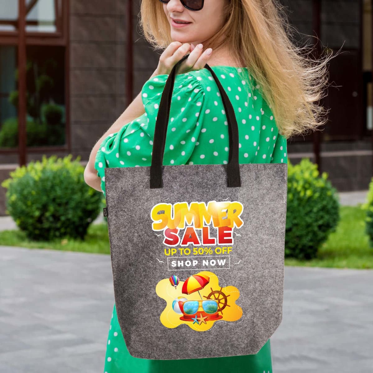 A woman in a green polka dot dress stands outdoors holding a gray tote bag. The bag is printed with a colorful Summer Sale design, advertising up to 50% off. The background shows a building with large windows.