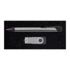 A silver pen with a black grip and a USB flash drive with a swivel cover are placed on a black textured surface. The pen is positioned horizontally above the USB drive.