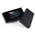 A black charging cable neatly coiled inside an open black box with a lid beside it. The cable has a USB connector at one end and a metallic tip at the other. The interior of the box is lined with soft, black material.