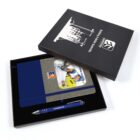 An open black box displays a gray notebook with a construction-themed cover inside, featuring workers wearing hard hats. Below the notebook is a blue pen labeled CAMCHAD. The box lid features a white building graphic and the text Building the Future.