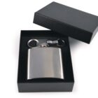 A stainless steel hip flask, bottle opener, and keyring are neatly presented in a black gift box with the lid partially open. The items are nestled in black foam, highlighting their sleek and polished design.