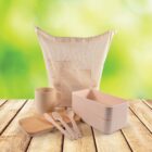 A set of eco-friendly dining items on a wooden surface. Includes a reusable bag, stacked lunchboxes, a cup, a wooden plate, and cutlery. The background is a blurred green and yellow, giving a natural vibe.