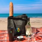 A beach picnic setup with a black tote bag containing a baguette. Nearby are two wine glasses, a wine bottle, a black tumbler, and a small round container, all set on a red plaid blanket with the ocean and sky in the background.