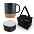 A black mug with a cork base, a black zippered lunch bag with a strap, and a round black portable speaker with a silver top, all set against a white background.