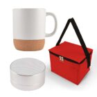 A white mug with a cork base, a small cylindrical silver speaker, and a red insulated lunch bag with a black handle and zippers are displayed on a white background.