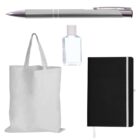 A collection of items against a white background, including a silver pen, a small clear bottle with liquid, a plain white tote bag, and a black notebook with a white elastic band and ribbon bookmark.
