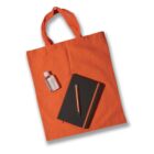 An orange tote bag lies flat with a black notebook and an orange pen resting on top. A small bottle of hand sanitizer is placed beside the notebook.