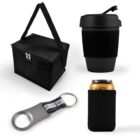 A collection of black travel accessories including a lunch bag with a zipper and strap, a reusable coffee cup with a lid, a metal bottle opener with a keychain ring, and a can holder with a black cover.