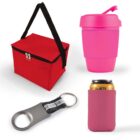 A red insulated lunch bag with a black strap, a pink reusable cup with a lid, a metal bottle opener with a black keyring strap, and a pink can cooler with a can inside are displayed against a white background.