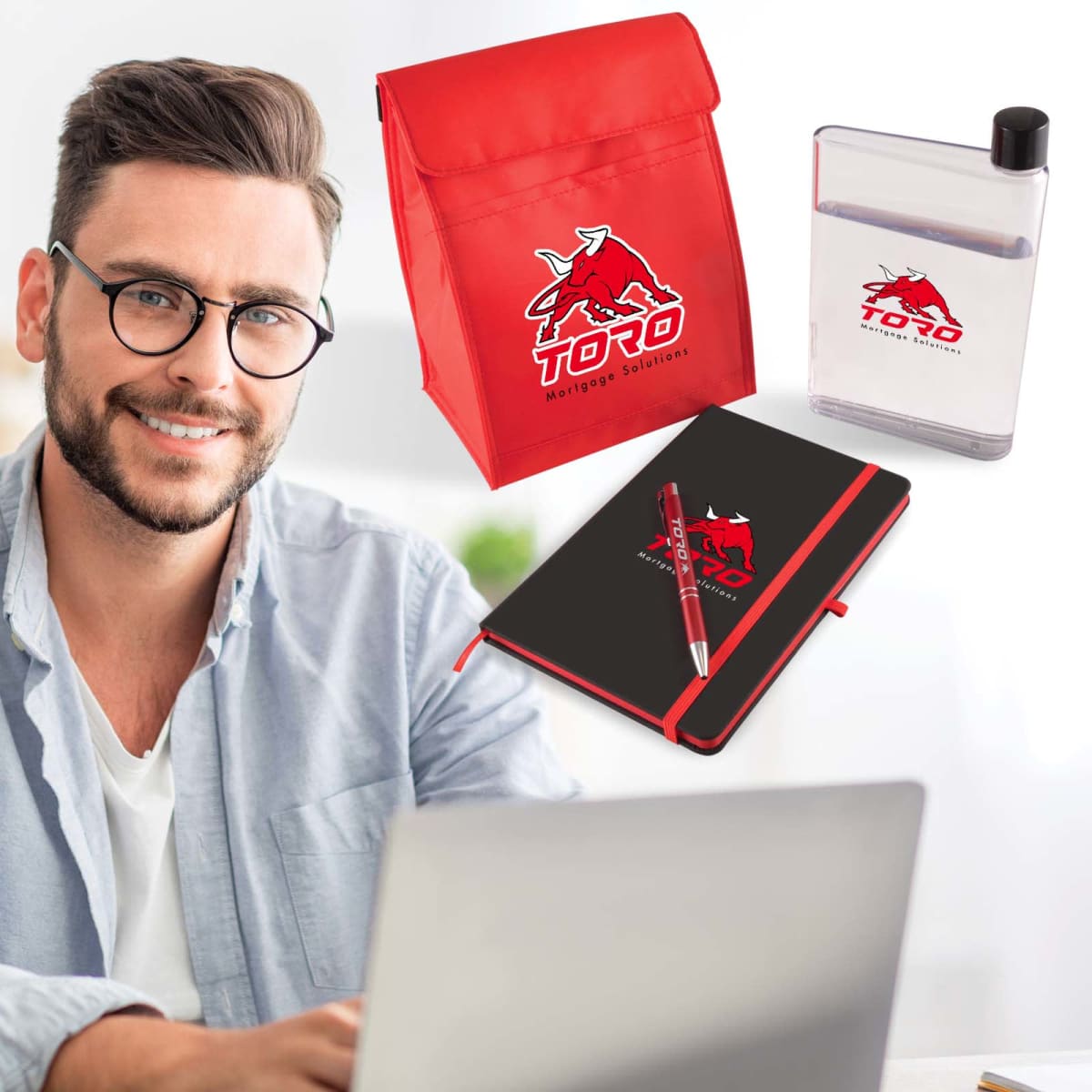 A person with glasses and a beard sits at a desk with a laptop. Nearby, branded items including a red lunch bag, notebook with a pen, and a clear water bottle, all featuring a red bull logo and Toro Mortgage Solutions text.