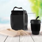 Reusable lunch set on a wooden surface includes a black insulated bag, a clear water bottle, a black travel mug, a beige compartmentalized container, utensils. Background is a blurred nature scene with green leaves and sky.